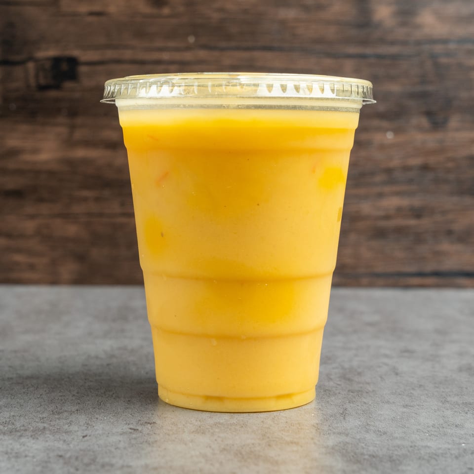 Bright yellow-orange beverage in a clear lidded plastic cup on a gray counter against a rustic wooden background.