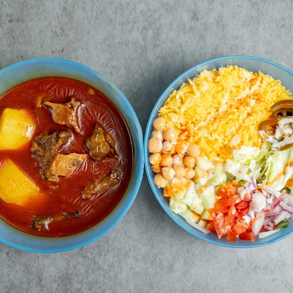 Two blue bowls hold red meat stew with potatoes and yellow rice served alongside chickpeas, salad, jalapeños, and creamy dressing.