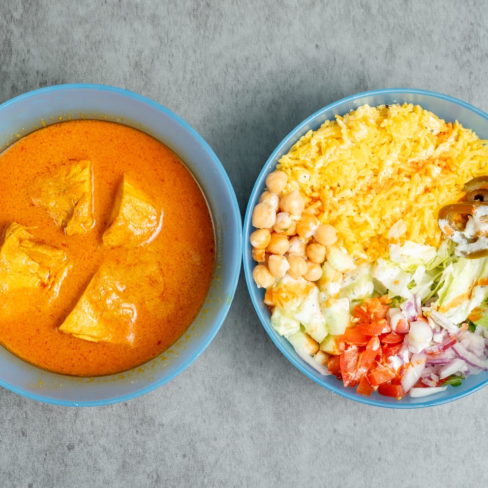 Bowls of creamy orange chicken curry and yellow rice with chickpeas, salad, pickles, and dressing on a gray surface.