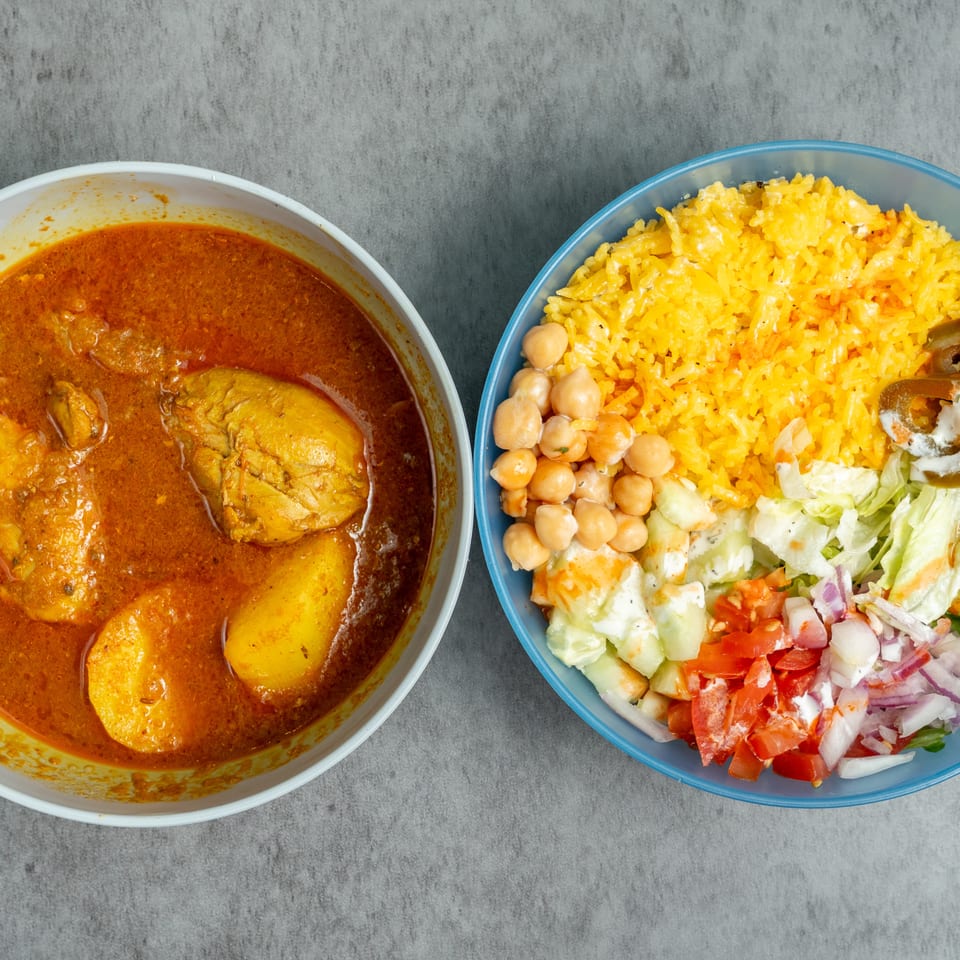 Bowls of chicken curry, yellow rice, chickpeas, jalapeños, and fresh salad arranged on a gray surface.