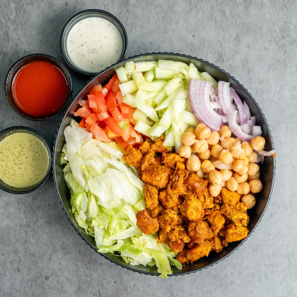 Overhead view of a chopped salad with lettuce, tomatoes, cucumber, red onion, chickpeas, crispy chicken, and three dipping sauces.